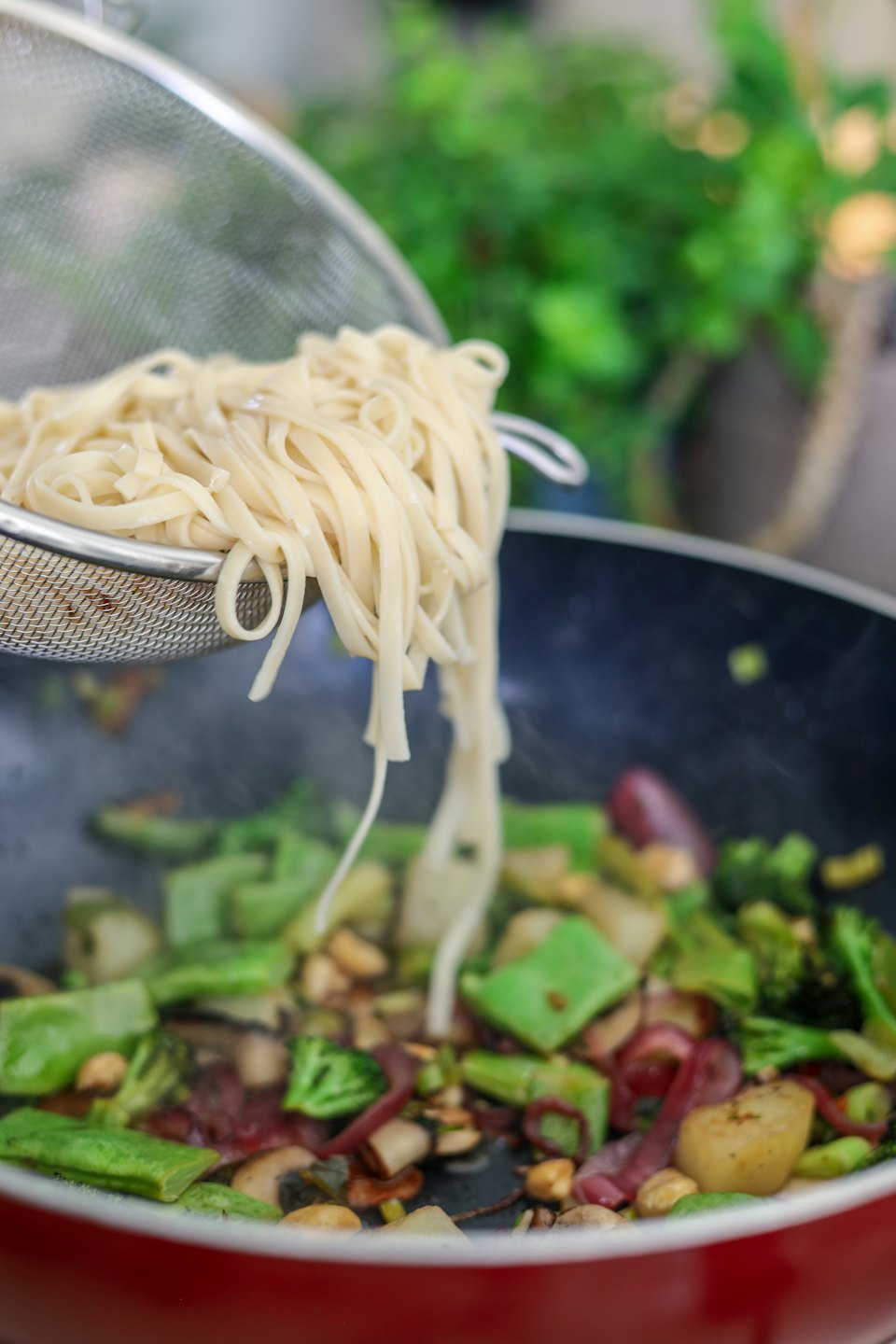 Biały makaron wsypywany do kolorowego stir-fry z warzywami w woku z czerwonym brzegiem.